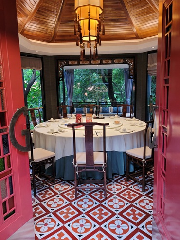 Un pavillon traditionnel chinois pour réunir les membres de la communauté autour d'un repas. La scène se déroule dans une pièce aux couleurs dominantes rouges. Au centre, une table ronde avec son plateau circulaire tournant à la présentation soignée. Une nappe blanche posée sur une table entièrement bordée d'une jupe bleue jusqu'au sol. Un luminaire traditionnel chinois avec sa lumière jaune tamisée fixé sur un plafond en dôme polygonal en bois forment un cercle protecteur parfait. L'arrière-plan est la grande baie vitrée du pavillon, ornée de tentures bleues, donnant sur un petit lac avec sa verdure luxuriante. Un canapé aux coussins bleus permet à quelques convives de trouver un espace isolé pour des échanges plus discrets. Un réalisme photographique raffiné souligne l'entrée dans un lieu traditionnel chinois fort en symbolique, avec une ambiance chaleureuse et feutrée qui favorise les rencontres de qualité et en toute de confiance.